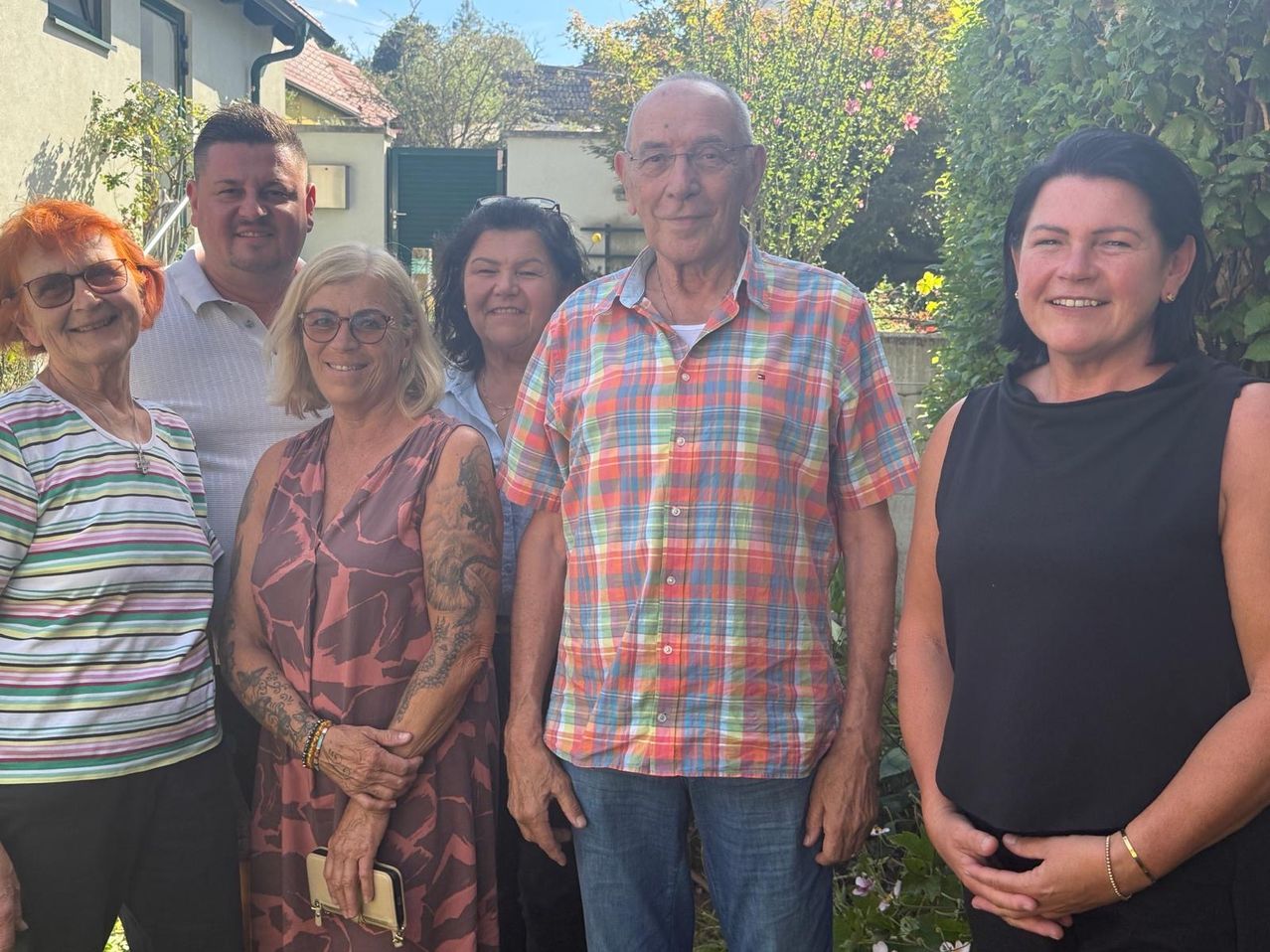 A group of six people are standing together in a garden, smiling for a photo. The man in the center wears a colorful plaid shirt, while the woman next to him has tattoos on her arms.