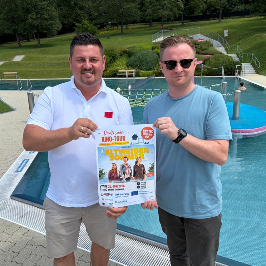 Zwei Männer stehen am Pool und halten ein Plakat für die Altweibersommer Kino-Tour. Das Plakat zeigt ein Bild von zwei Personen.