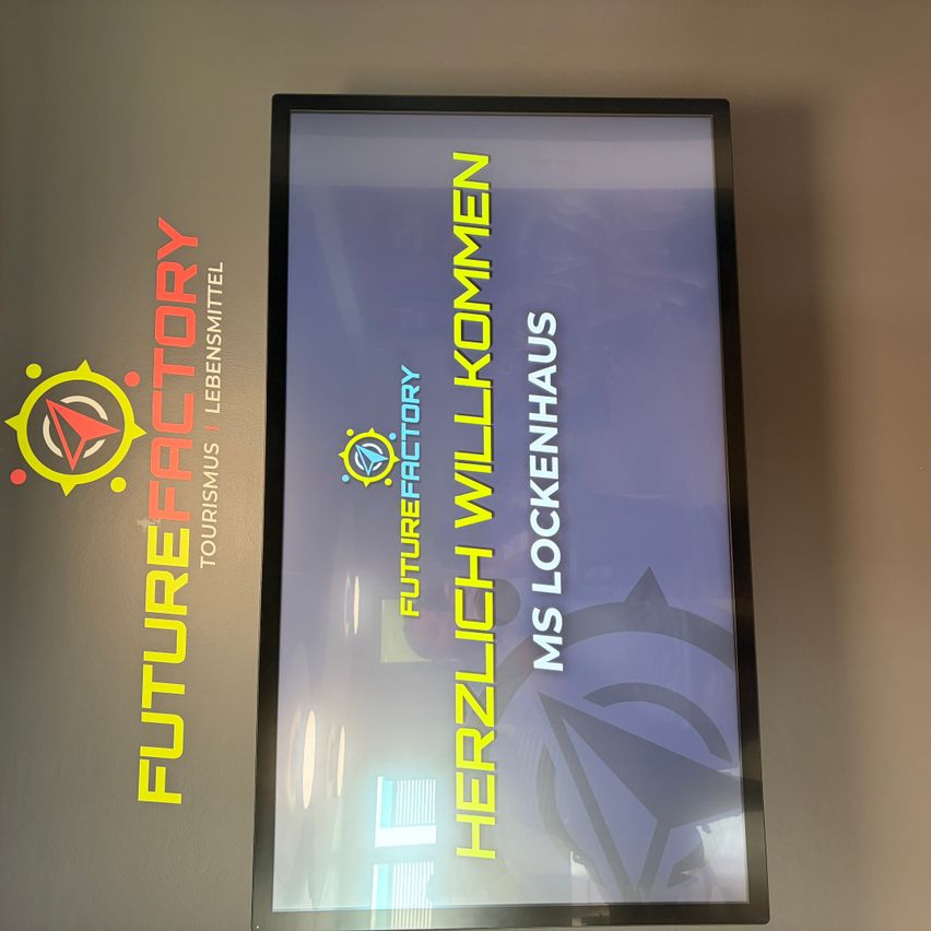 Ein Monitor zeigt eine Willkommensnachricht auf Deutsch an. Es steht Herzlich Willkommen und Ms. Lockenhaus. Der Monitor ist an einer Wand angebracht.