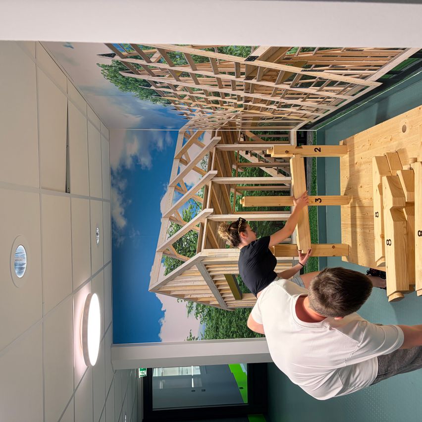 Zwei Personen arbeiten an einer Holzstruktur in einem Raum mit einer Wandmalerei eines Baumhauses an der Decke.