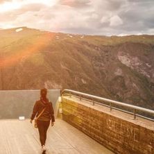 Eine Frau geht auf einer hölzernen Brücke mit Metallgeländer und trägt eine Tasche. Die Brücke ist erhöht und bietet einen Blick auf einen steilen, felsigen Berg mit bewölktem Himmel oben.