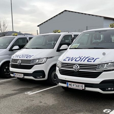 Drei weiße Volkswagen-Transporter mit der Aufschrift 'zwolfer' sind auf einem Parkplatz geparkt. Die Transporter haben Taxischilder oben und Nummernschilder. Dahinter befindet sich ein Gebäude mit Zaun.