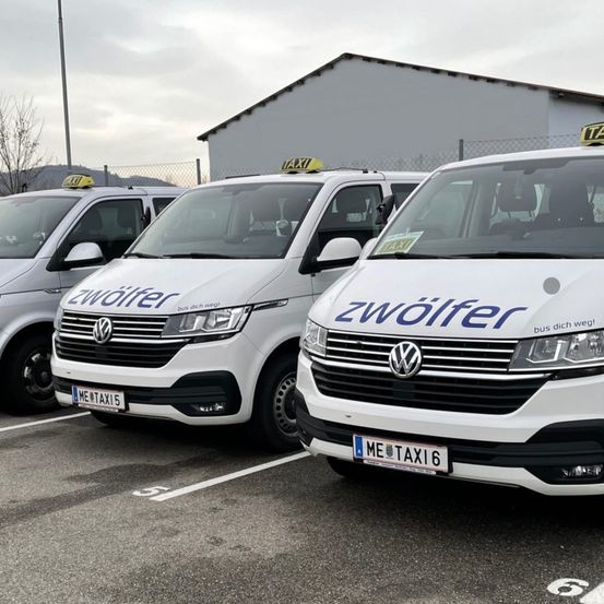 Drei weiße Volkswagen-Transporter mit der Aufschrift 'zwolfer' sind auf einem Parkplatz geparkt. Die Transporter haben Taxischilder oben und Nummernschilder. Dahinter befindet sich ein Gebäude mit Zaun.