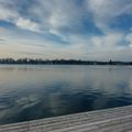 Ein ruhiger See mit ruhigen Gewässern unter einem bewölkten Himmel, ein hölzerner Steg, der ins Wasser ragt. Bäume und Gebäude säumen den Horizont in der Ferne.