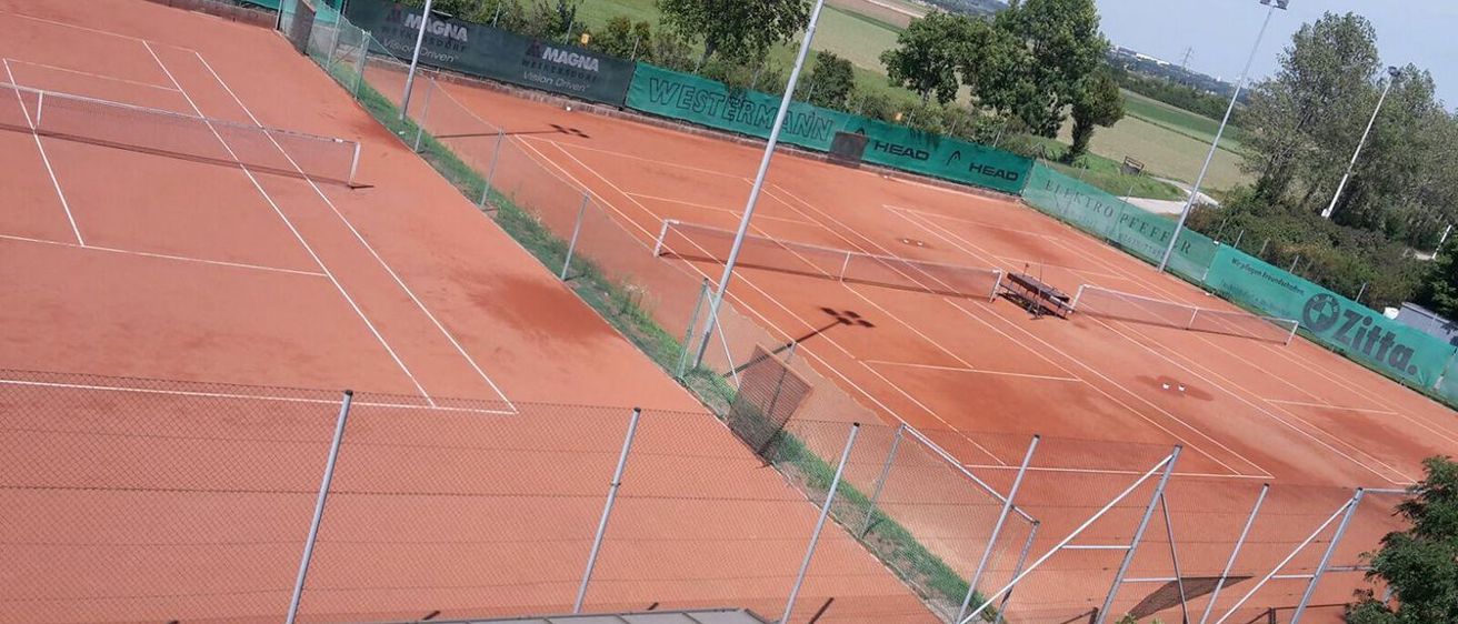 Ein Blick von oben auf einen Tennisplatz mit rotem Sandboden und Metallzäunen, mit Sponsorenbannern wie MAGNA und WESTERNMANN, umgeben von viel Grün.