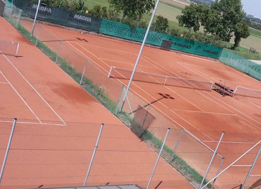 Ein Blick von oben auf einen Tennisplatz mit rotem Sandboden und Metallzäunen, mit Sponsorenbannern wie MAGNA und WESTERNMANN, umgeben von viel Grün.