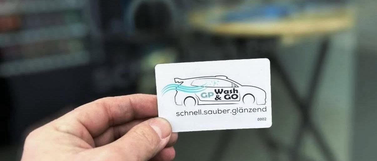 Eine Person hält eine weiße Visitenkarte mit dem Logo von GP Wash & Go, das einen Wagen und Wasserwellen zeigt, mit einem unscharfen Hintergrund eines Autowaschgeschäfts.