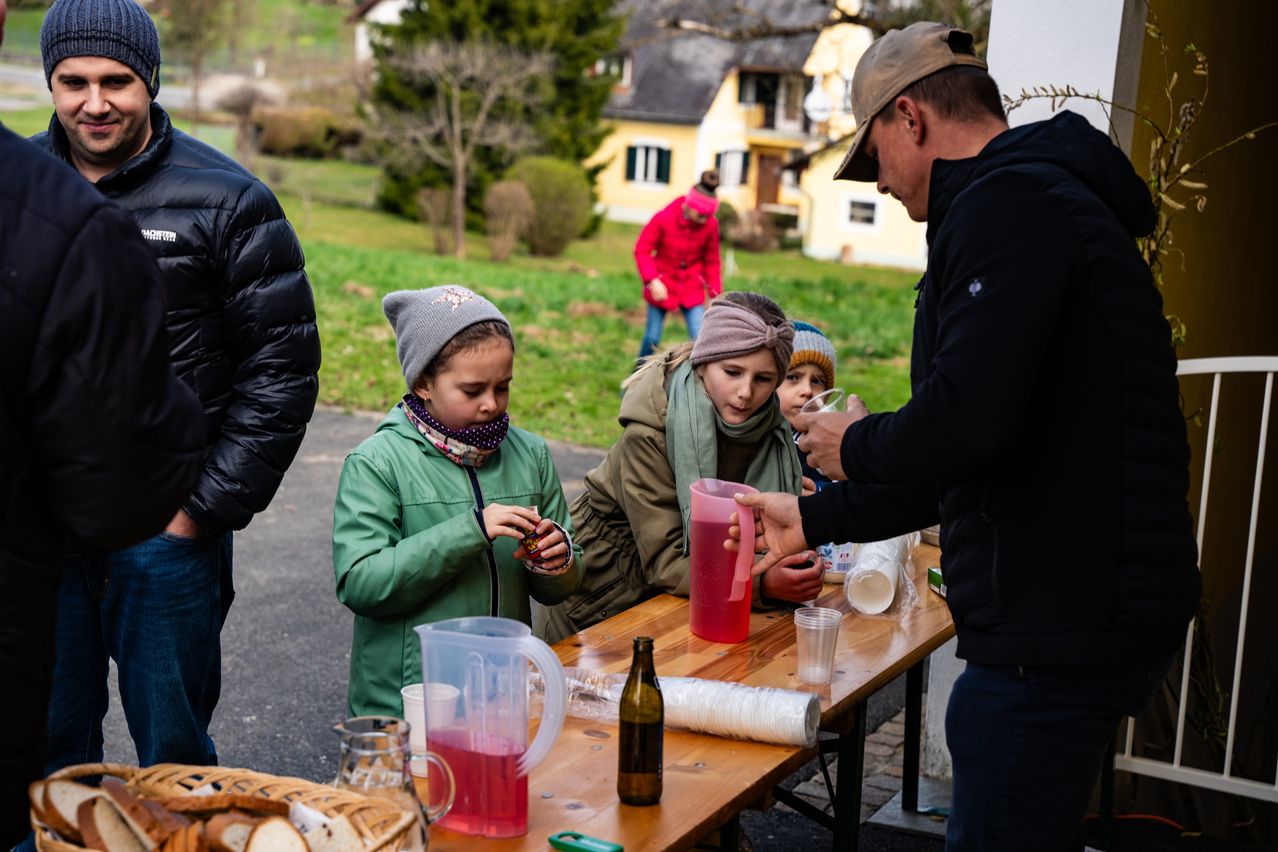 Eine Gruppe von Kindern und Erwachsenen versammelt sich um einen Tisch, ein Mann serviert ihnen Getränke und ein Haus ist im Hintergrund.