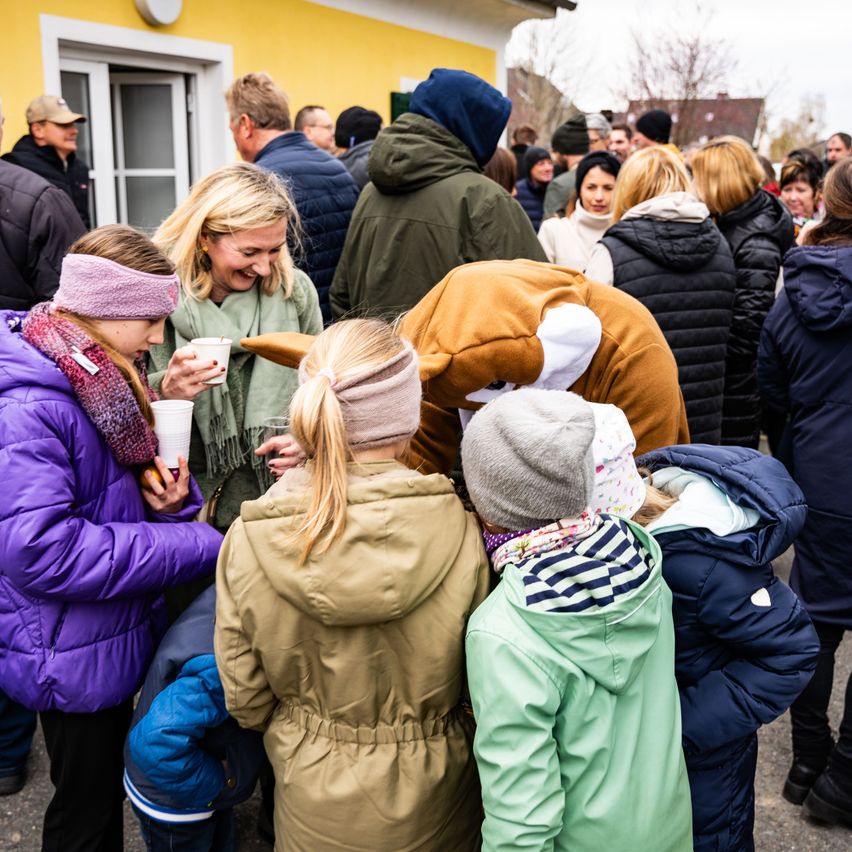 Eine Menschenmenge versammelt sich draußen; Frauen und Kinder halten Tassen und lächeln. Eine Frau in einem grünen Mantel steht zwischen ihnen, und ein Kind in einem Fuchskostüm beugt sich vor. Im Hintergrund befindet sich ein Gebäude mit einer weißen Tür.