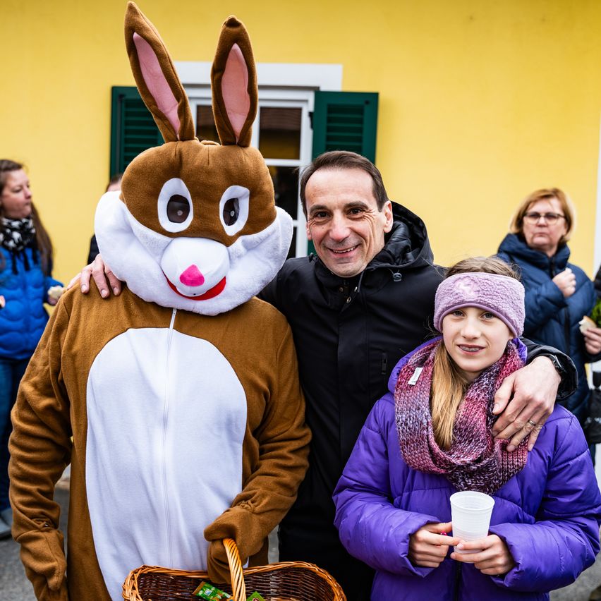 Ein Mann und ein Mädchen in Winterkleidung stehen mit einer Person in einem Kaninchenkostüm, halten einen Korb und einen Becher. Hinter ihnen steht eine weitere Frau an einer gelben Wand.