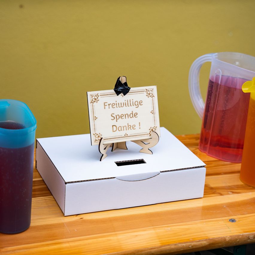 Eine weiße Box mit einem Schild, auf dem 'Freiwillige Spende Danke!' steht, steht auf einem Holztisch mit zwei Krügen mit roter Flüssigkeit.