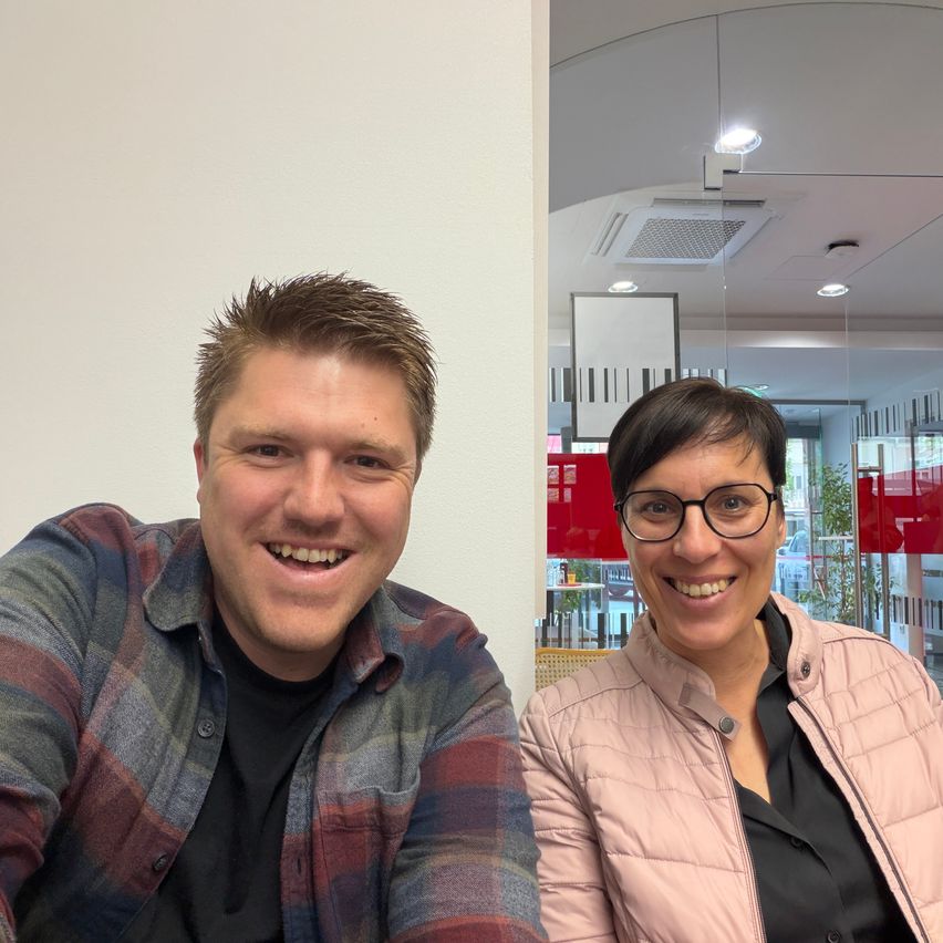 A man and a woman are smiling and posing for a picture in an indoor setting. The man is wearing a plaid shirt.