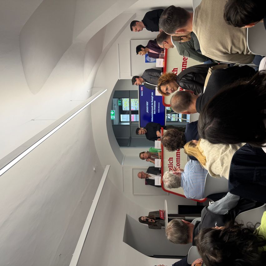 A crowd of people are gathered in a room, possibly for a meeting. Some are standing while others are sitting. A banner with a logo and text is displayed in the center.