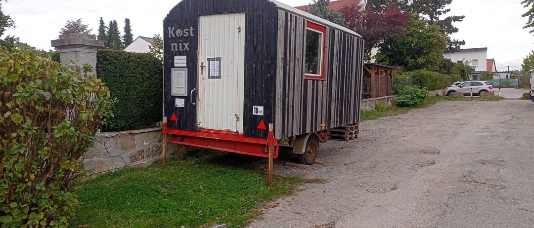 Ein kleiner, schwarz-weißer Wohnwagen steht am Straßenrand, mit rotem Zierrat und weißer Tür. Ein Schild zeigt 'Kest nix' an.