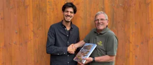 Zwei Männer stehen vor einer Holzwand, einer hält ein Buch und lächelt, der andere schüttelt ihm die Hand.