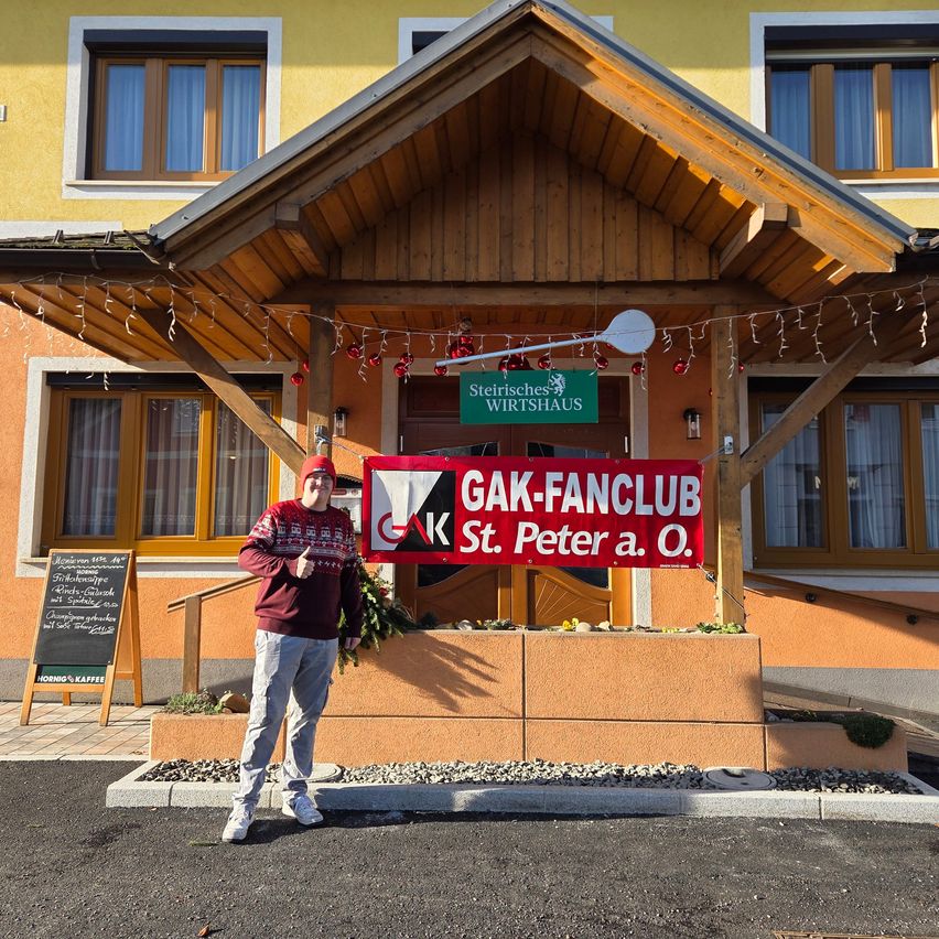 A person stands in front of a building with a sign saying 'GAK-FANCLUB St. Peter a. O.' The building has wooden walls and yellow paint.