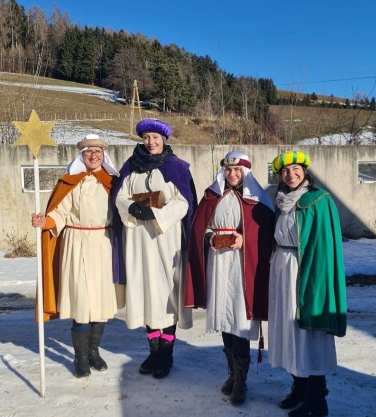 Vier Personen in traditionellen Kostümen stehen im Schnee, wahrscheinlich für ein Fest. Sie tragen verschiedenfarbige Umhänge und Hüte. Eine Frau hält einen Stern. Hinter ihnen ist eine Betonmauer mit einem Fenster zu sehen, mit Bäumen und Bergen im Hintergrund.