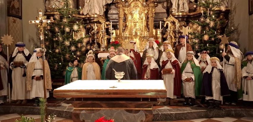 Eine Gruppe von Menschen in festlichen Kostümen und Hüten steht vor einem Altar. Der Altar ist mit einem weißen Tuch und einem Kelch geschmückt. Dahinter stehen Weihnachtsbäume und Statuen.