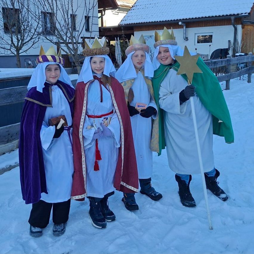 Vier Kinder in farbenfrohen Kostümen stehen im Schnee. Sie tragen Kronen und halten Sternenstäbe in der Hand, während sie vor einem verschneiten Haus für ein Foto posieren.