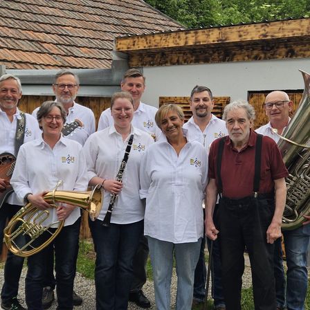 Eine Gruppe von Musikern posiert für ein Foto vor einem Haus. Sie halten Musikinstrumente wie Tuba, Klarinette und Gitarre.
