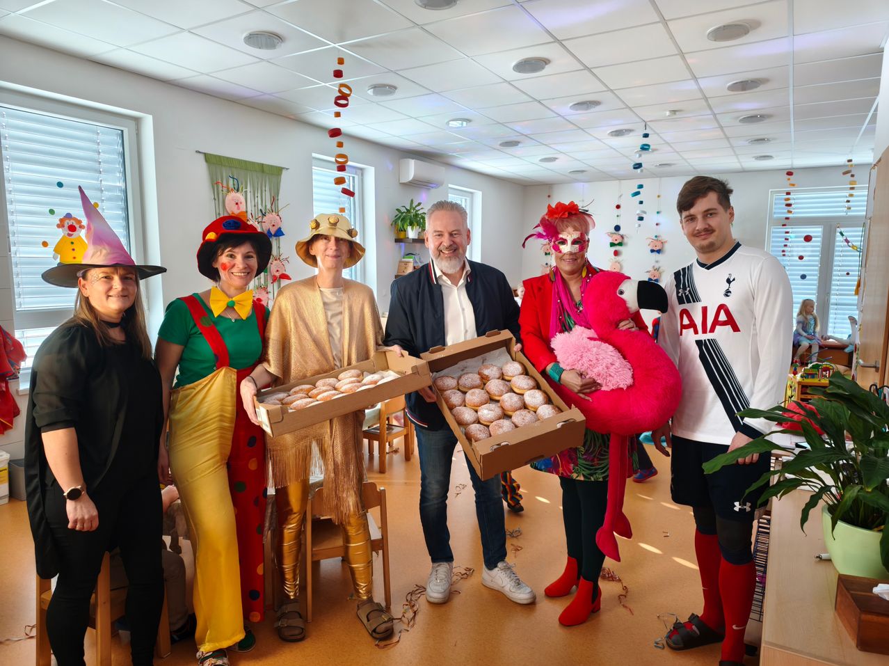 Eine Gruppe von Menschen in bunten Kostümen steht in einem Raum und hält Schachteln mit Donuts. Eine Person hält einen großen rosa Flamingo-Plüsch.