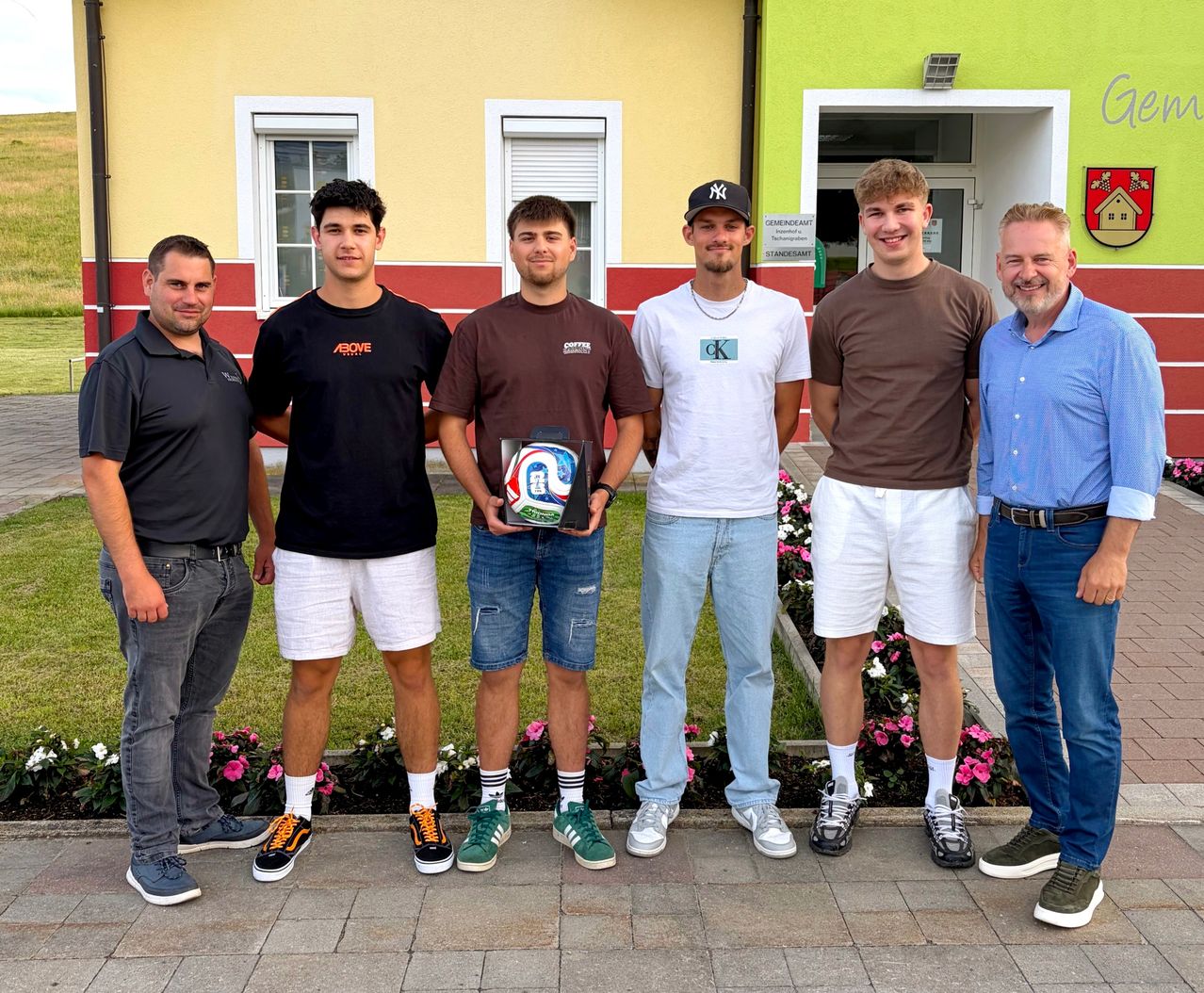 Sechs Männer stehen lächelnd in einer Reihe vor einem Gebäude mit Blumen. Ein Mann hält einen Fußballpokal.