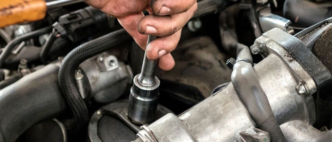 Eine Person arbeitet mit einem Schraubenschlüssel an einem Fahrzeugmotor und konzentriert sich auf eine metallische Komponente. Der Motor ist von verschiedenen metallischen Teilen und Drähten umgeben.