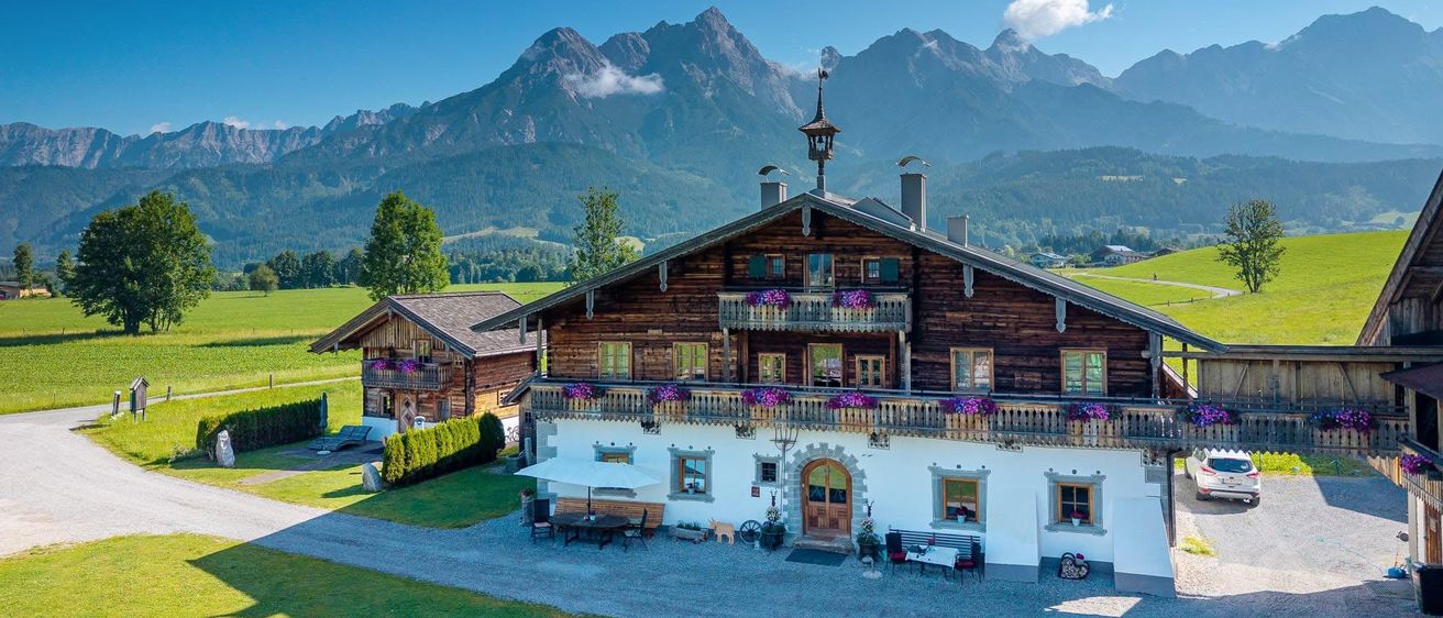 Ein Luftbild eines traditionellen Holzchalets mit großer Terrasse und Blick auf die Berge. Das Chalet ist von einem üppigen grünen Feld umgeben.