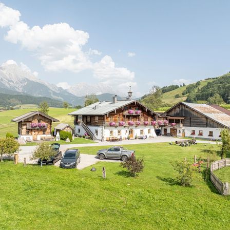 Luftaufnahme eines Bauernhofs mit mehreren Gebäuden, einem großen zentralen Haus mit Blumen auf dem Balkon und zwei kleineren Häusern. Drei Autos sind vor dem Hof geparkt, mit Bergen im Hintergrund.