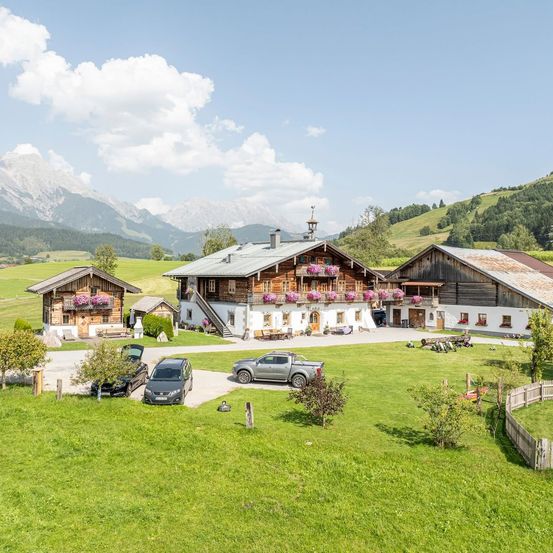 Luftaufnahme eines Bauernhofs mit mehreren Gebäuden, einem großen zentralen Haus mit Blumen auf dem Balkon und zwei kleineren Häusern. Drei Autos sind vor dem Hof geparkt, mit Bergen im Hintergrund.