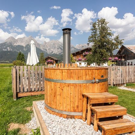 Eine Holzfasssauna in einem Grasbereich mit Bergen und Häusern im Hintergrund. In der Nähe befinden sich Holzstufen und ein Zaun.