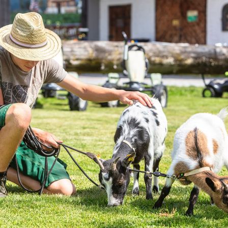 Ein junger Mann mit Strohhut kniet auf dem Rasen und füttert zwei Ziegen mit einem Seil. Die Ziegen sind schwarz-weiß und braun-weiß. Im Hintergrund befinden sich ein Baumstamm, ein Fahrzeug und ein Gebäude.