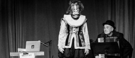 Ein Schwarz-Weiß-Foto eines maskierten Menschen, der auf der Bühne steht und einen Akkordeon spielenden sitzenden Mann. Hinter dem Darsteller steht ein Tisch mit einem Laptop und einem Schild mit der Aufschrift 'Gesucht' und zwei Fotos.