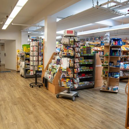 Der Innenraum eines Geschäfts mit Holzboden zeigt verschiedene Produkte auf Regalen. Es gibt bunte Bücher, Zeitschriften und andere Artikel. Der Laden hat Deckenleuchten und eine weiße Säule in der Mitte.