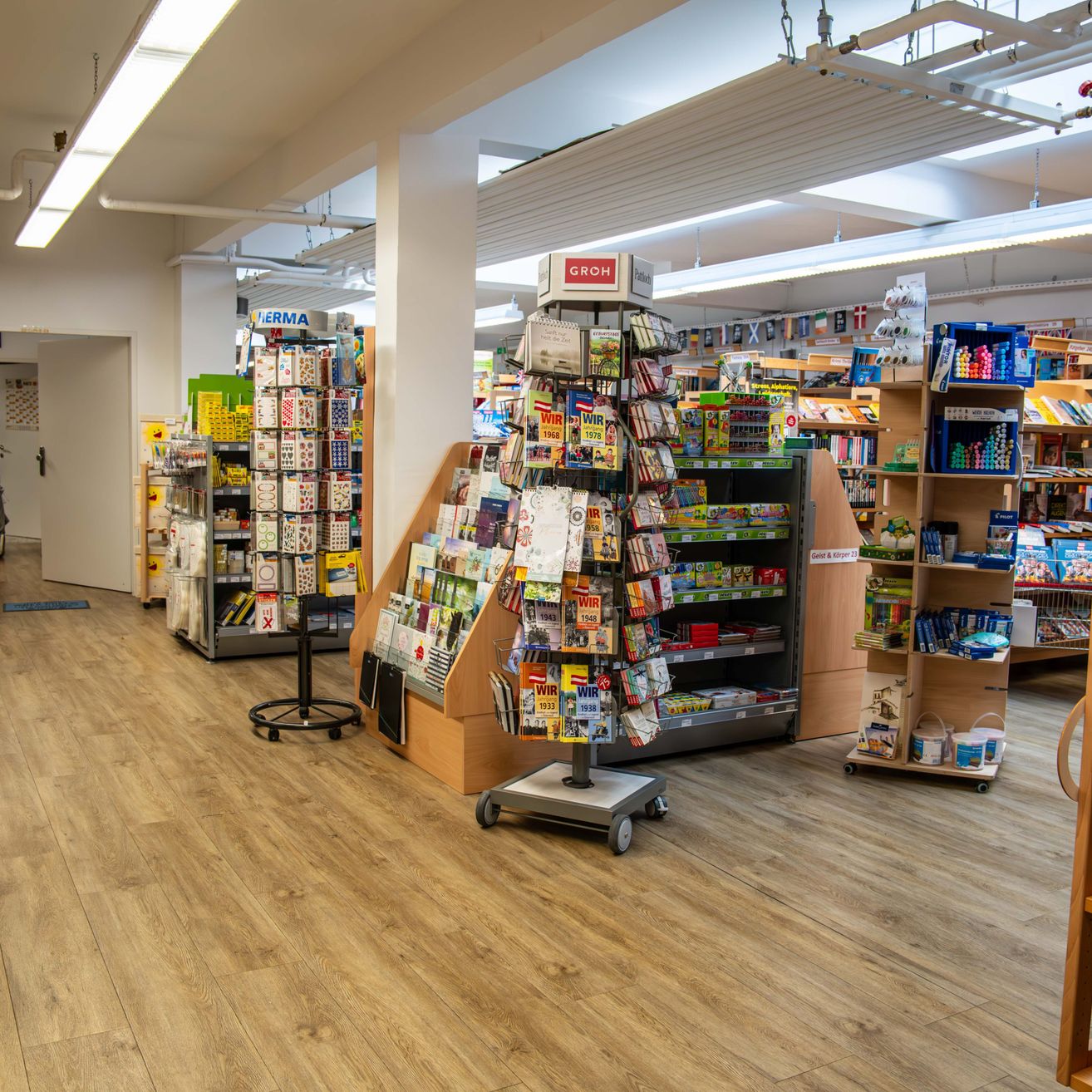 Der Innenraum eines Geschäfts mit Holzboden zeigt verschiedene Produkte auf Regalen. Es gibt bunte Bücher, Zeitschriften und andere Artikel. Der Laden hat Deckenleuchten und eine weiße Säule in der Mitte.