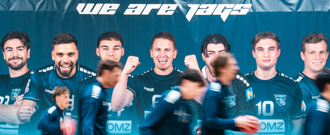 Fünf Männer in blauen Trikots stehen vor einem Banner mit der Aufschrift 'We are Jags'. Der Mann in der Mitte hebt die Faust.