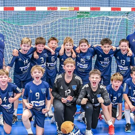 Eine Gruppe junger Jungen in blauen Trikots mit Nummern von 1 bis 16 posiert für ein Foto vor einem Handballtor. Sie haben den Mund offen und scheinen zu schreien.