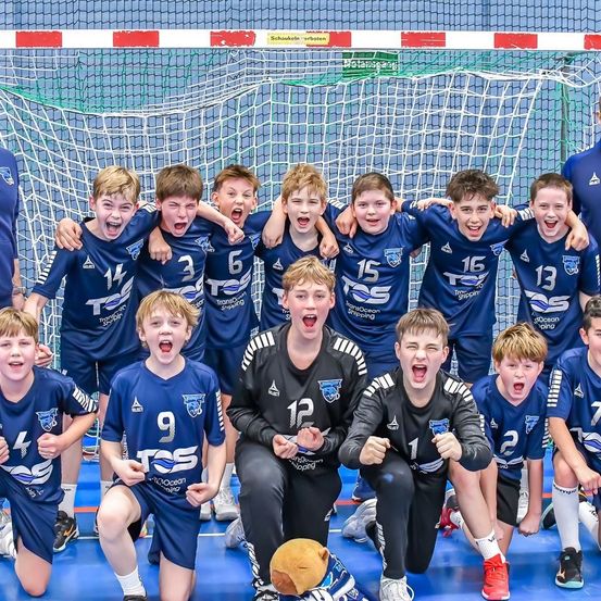 Eine Gruppe junger Jungen in blauen Trikots mit Nummern von 1 bis 16 posiert für ein Foto vor einem Handballtor. Sie haben den Mund offen und scheinen zu schreien.