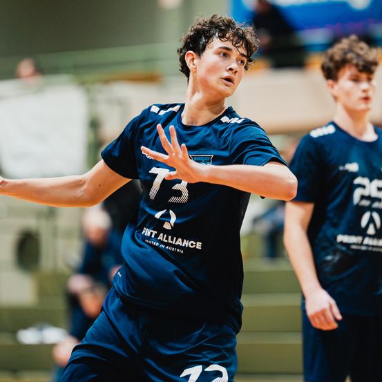 Ein junger Mann in einem blauen Sporttrikot mit der Nummer 73 spielt ein Spiel, wahrscheinlich Basketball, mit einem anderen Spieler. Sie befinden sich in einer Sporthalle.