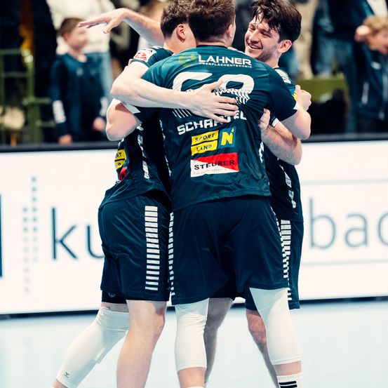 Eine Gruppe von Handballspielern in Uniform umarmt sich zur Feier. Einer trägt die Nummer 29. Das Publikum schaut von einer Barriere aus zu.