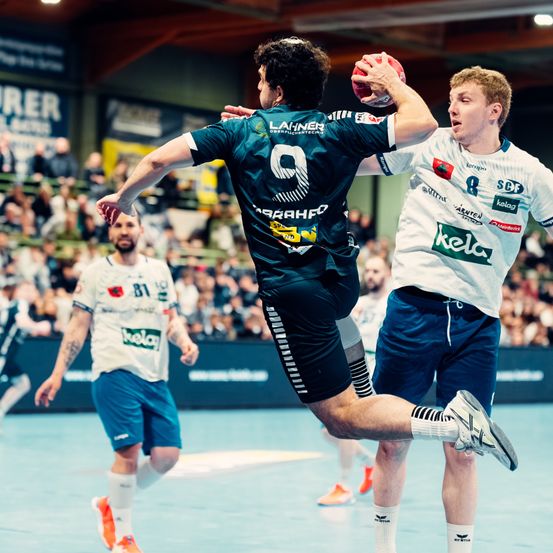 Zwei Handballspieler sind in ein Spiel verwickelt, einer in einem blauen Trikot mit der Nummer 9, der andere in Weiß. Sie sind von Teamkollegen umgeben, Zuschauer beobachten von den Tribünen.