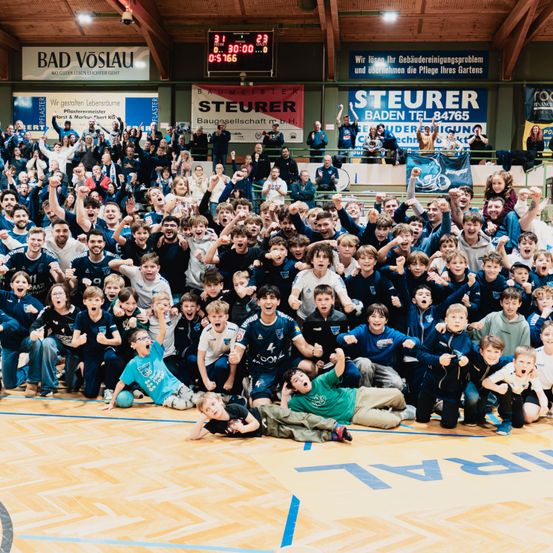 Eine Gruppe von Kindern und Erwachsenen jubelt in einer Sporthalle, wahrscheinlich nach einem Sportereignis. Einige tragen blaue Trikots, während andere Freizeitkleidung tragen. Banner mit Text hängen an den Wänden.