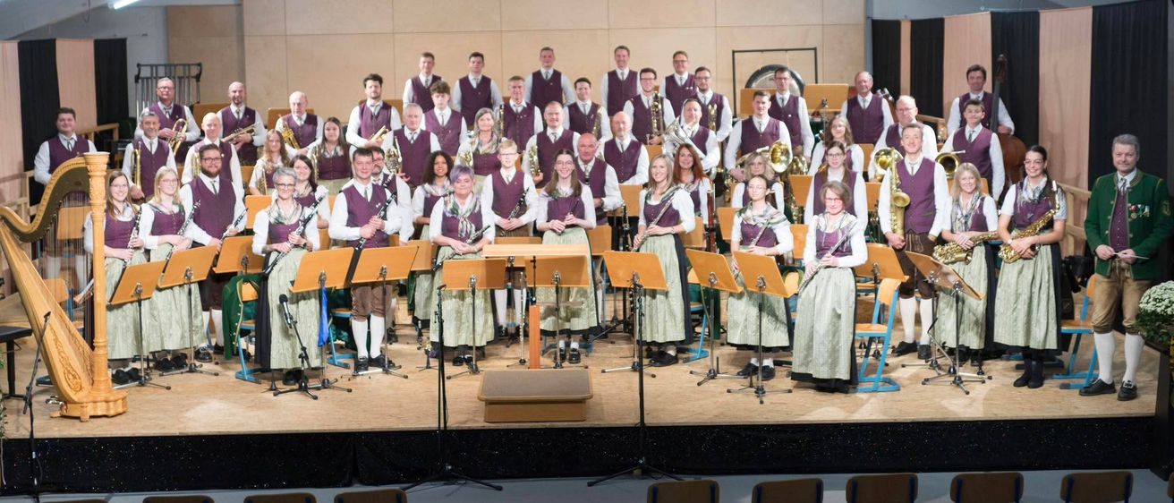 Ein großes Ensemble von Musikern in einheitlichen Kostümen, die mit verschiedenen Instrumenten wie Klarinetten und Trompeten auf der Bühne stehen, vor leeren Stühlen.