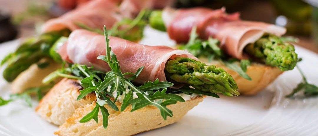Ein Teller zeigt getoastetes Brot, das mit in Prosciutto eingewickeltem Spargel garniert ist, dekoriert mit Rucola.