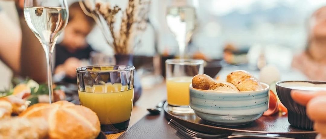 Ein Frühstückstisch mit Gläsern Orangensaft, Croissants in einer Schüssel, Brot und Besteck. Eine Vase mit getrockneten Blumen und einer verschwommenen Person im Hintergrund.