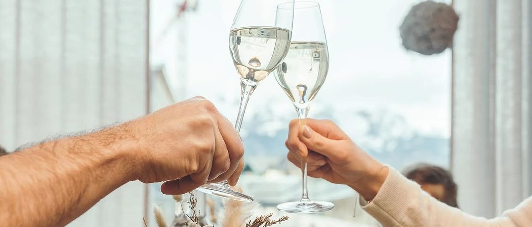 Zwei Personen halten Weinglaser, möglicherweise zum Anstoßen, in einer Innenumgebung mit einem Fenster dahinter.
