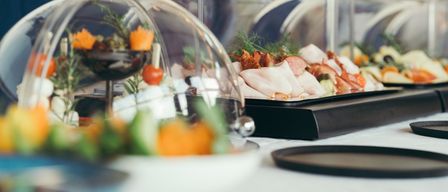 Ein Tisch mit mehreren durchsichtigen Glaskuppeln und Tabletts mit geschnittenem Fleisch, Gemüse und Garnierungen. Jede Kuppel steht auf einer schwarzen Basis mit einem kleinen dekorativen Teller in der Nähe.