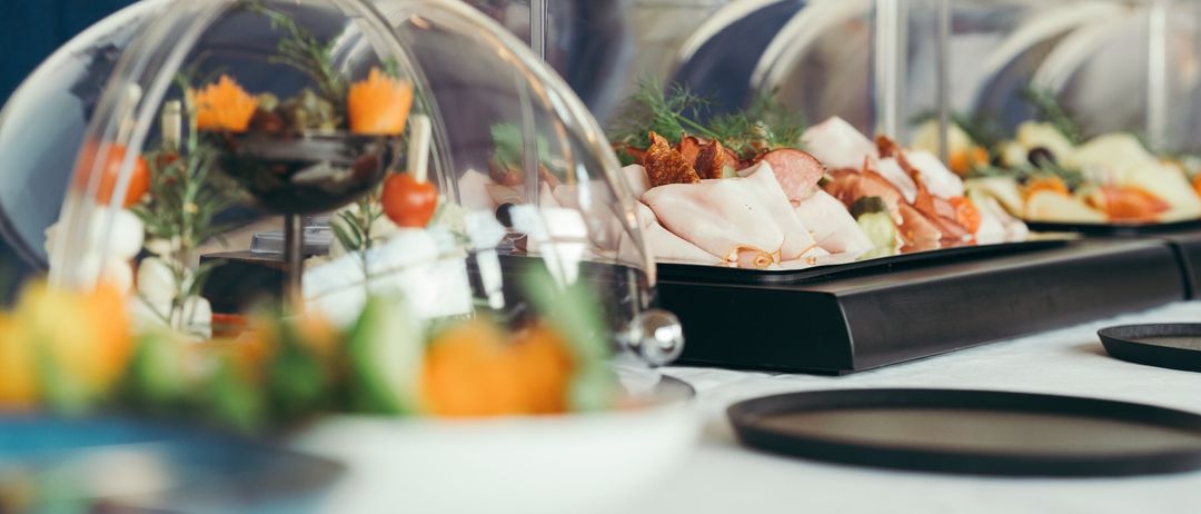 Ein Tisch mit mehreren durchsichtigen Glaskuppeln und Tabletts mit geschnittenem Fleisch, Gemüse und Garnierungen. Jede Kuppel steht auf einer schwarzen Basis mit einem kleinen dekorativen Teller in der Nähe.