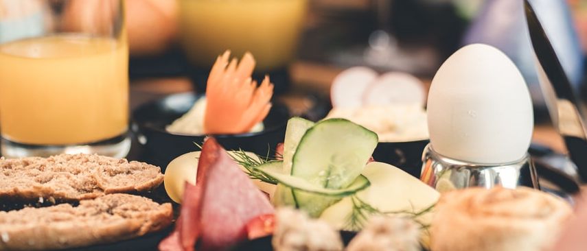 Eine Person sitzt an einem Tisch mit einer Auswahl an Frühstücksspeisen, darunter Brot, Eier und Gemüse. Auf dem Tisch stehen auch Gläser Orangensaft und Wein.