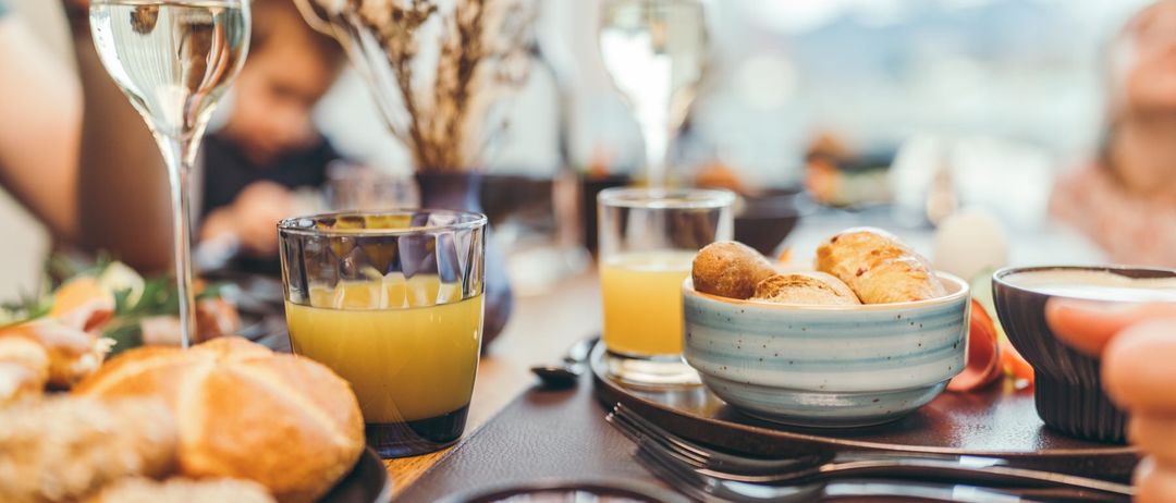 Ein Tisch mit einer Reihe von Gläsern, Schüsseln und Tellern mit Brot und Croissants. Eine Person sitzt im Hintergrund, und eine Vase mit getrockneten Blumen steht auf dem Tisch.